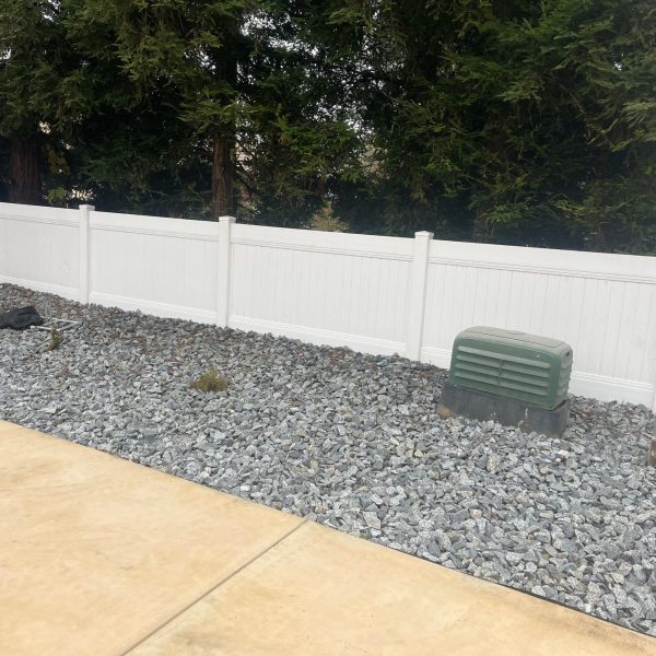 A Neatly Landscaped Area With Gray Rocks A White Fence And Evergreen Trees. A Green Utility Box On One Side Adds Contrast Against The Neutra Scaled